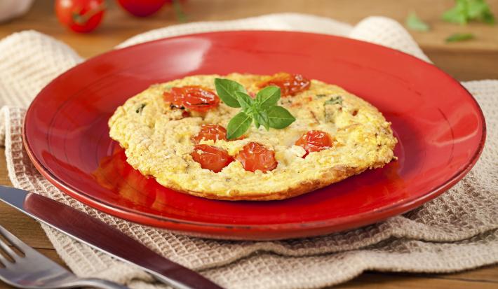 Fotografia em tons de vermelho em uma bancada de madeira, um pano bege, um prato vermelho redondo com a omelete com tomates-cereja em cima dele. Ao fundo, tomatinhos para decorar e ao lado, garfo e faca.