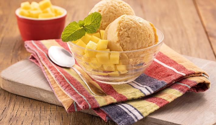 Foto da receita de sorbet de manga servida com uma folha de hortelã em uma taça de vidro sobre um pano colorido listrado. Ao lado há uma colher prateada como decoração, bem como um recipiente vermelho com mangas picadas ao fundo.