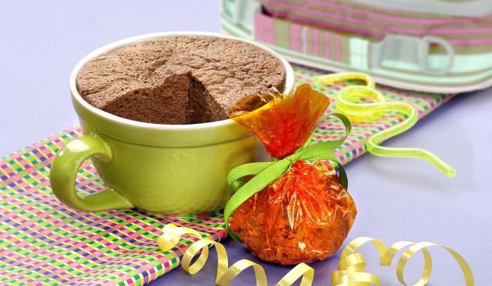 fotografia em tons de cinza, marrom e verde de uma bancada cinza vista de frente, contém um pano em tons de verde, rosa e roxo e uma xicara verde com bolo de chocolate dentro.
