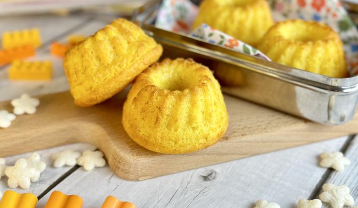 Fotografia em tons de laranja em uma bancada de madeira clara, uma lancheira ao meio com alguns bolinhos de milho dentro e em cima de uma tábua de madeira. Ao lado, brinquedos infantis.