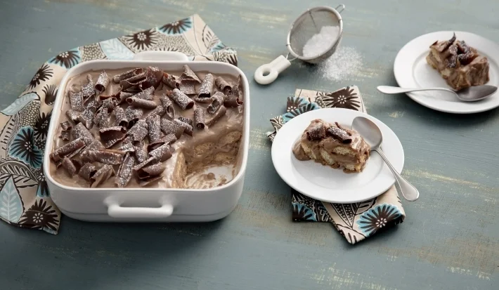 Travessa quadrada com pavê de chocolate decorado com raspas de chocolate e dois pratos com um pedaço.