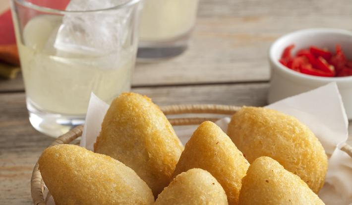 Fotografia em tons de dourado e grafite, com cestinha de vime ao centro contendo 6 coxinhas fritas, no lado direito potinho com pimentas vermelhas, ao alto dois copos com suco e gelo e um guardanapo vermelho e branco, tudo sobre bancada rústica.