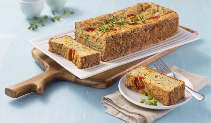Foto da receita de Tortinha de Legumes. Observa-se uma superfície azul clara, com uma tábua de madeira e, em cima, um prato branco retangular com a torta de legumes inteira, também retangular. Ao lado, um prato branco redondo com um pedaço da torta.