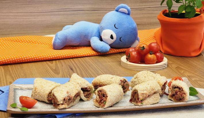 Fotografia de oito rolinhos de pão com atum e mucilon sobre um apoio de vidro retangular grande. No fundo tem um ursinho azul de mucilon deitado sobre um paninho laranja com bolinhas brancas, ao lado de alguns tomates cereja e um vasinho de plantas.