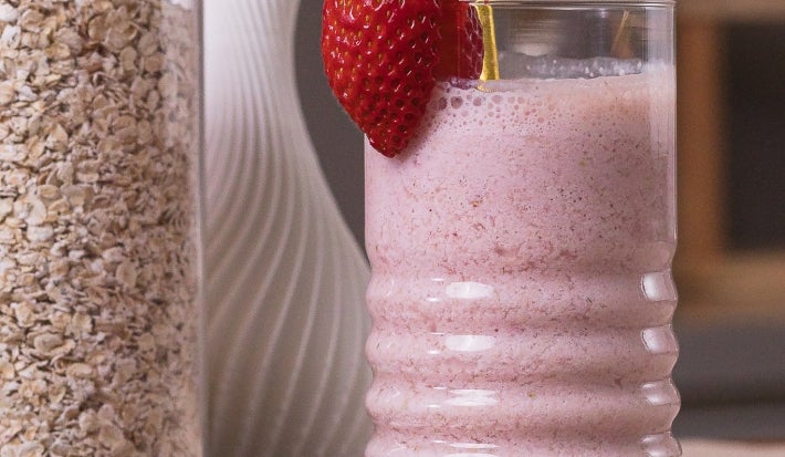 Smoothie pós-treino de morango em copo de vidro com canudo dourado e morango na borda, acompanhada por morangos frescos e pote de aveia.