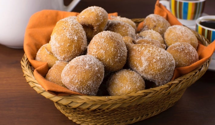 Em cima de uma mesa, cesta de palha com pano laranja com bolinhos de chuva, polvilhado com açúcar de confeiteiro.