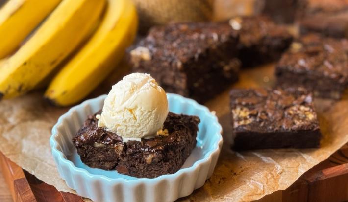 Fotografia de uma mini forma de vidro branca com um pedaço do brownie de banana com uma bola de sorvete de creme por cima. O recipiente está sobre uma folha de papel manteiga com mais pedaços do brownie e um cacho de bananas.