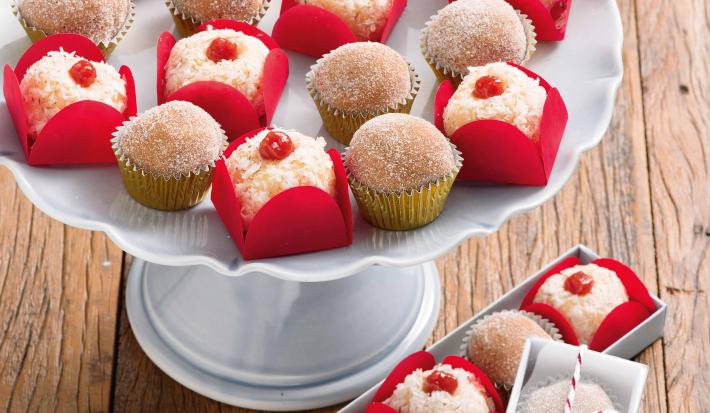 Fotografia em tons de vermelho em uma bancada de madeira com um suporte branco para bolo e vários brigadeiros de churros e com goiabada em cima dele dentro de forminhas vermelhas de docinhos.