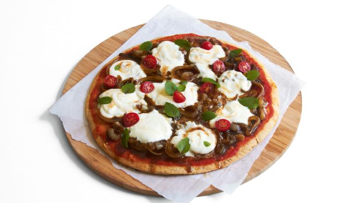 Ao centro, uma tábua de madeira marrom redonda com um pano branco e uma pizza com tomates, cebolas e iogurte por cima.