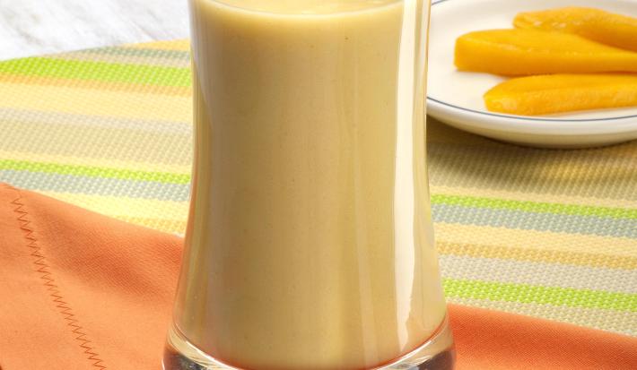 Fotografia em tons de laranja em uma bancada de madeira com uma toalha colorida listrada, um pano laranja e um copo de vidro alto com a vitamina de banana com cupuaçu. Ao fundo, um pratinho branco raso com fatias de manga.