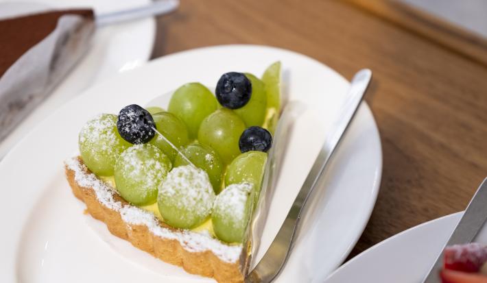 Fotografia de uma fatia de torta recheada com leite MOÇA e uvas verdes por cima, com três uvas roxas. Por cima da torta tem açúcar de confeiteiro polvilhado, e ao lado, um garfo sobre um prato branco, que está sobre uma mesa de madeira.