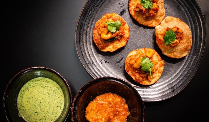 Fotografia vista de cima de um prato preto raso com quatro unidades de pani puri com grão de bico e coentro. Ao lado tem dois molhos em recipientes diferentes, um molho de iogurte e o outro, um molho de manga. Tudo está sobre uma mesa preta.