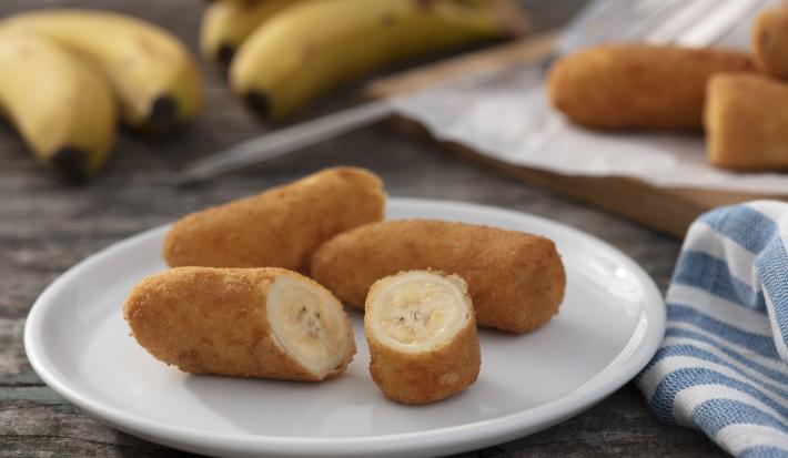 Imagem de um prato branco uma uma banana a milanesa cortada ao meio. Ao fundo, bananas e um paninho listrado branco com azul