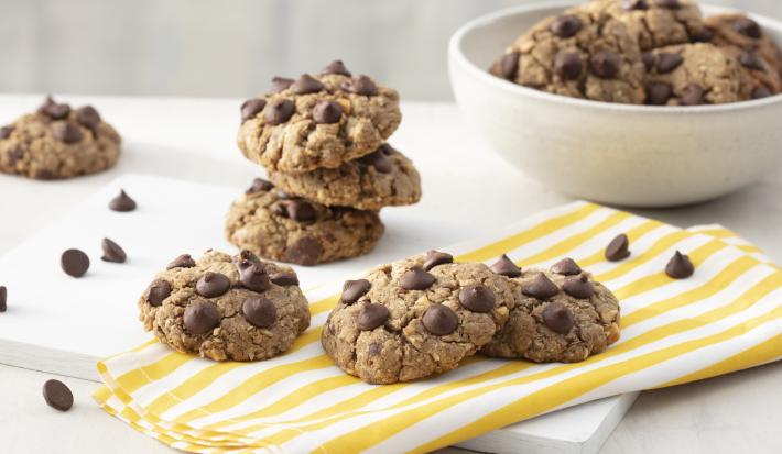 Fotografia em tons de branco e amarelo de uma bancada branca com uma tabua branca e um paninho listrado amarelo e branco sobre ele cookies. Ao fundo um recipiente redondo branco com cookies e gotas de chocolate espalhadas pela mesa.