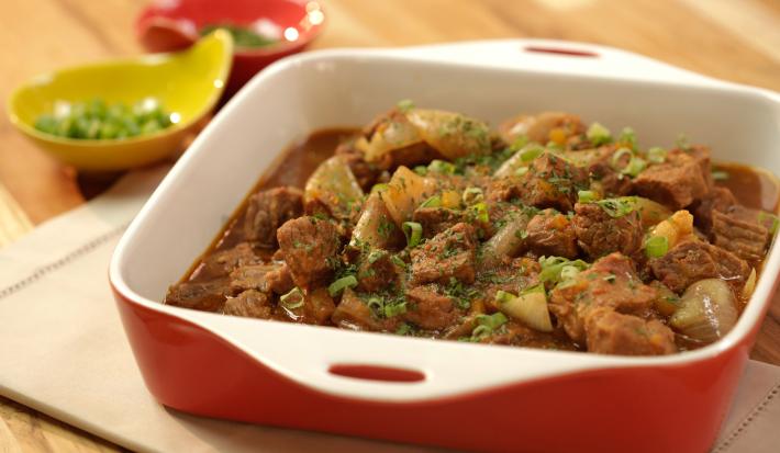Em uma mesa de madeira contém um pano branco e por cima um recipiente nas cores vermelho e branco, que contém cubos de carne. Ao lado dois potes na cor amarelo e vermelho.