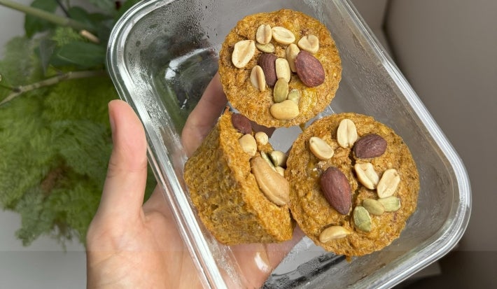 Fotografia em tons claros de uma mão segurando um recipiente de vidro pequeno e retangular. Dentro do recipiente tem três muffins de banana e aboora, decorados com castanhas.