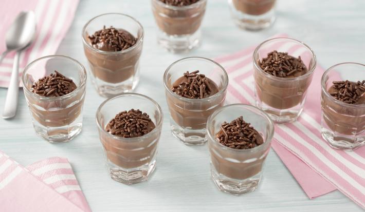 Fotografia em tons de azul e rosa claros de uma bancada azul clara com paninhos rosas com listras brancas, sobre eles nove copinhos de vidro redondos com brigadeiro de colher e granulados de chocolate.