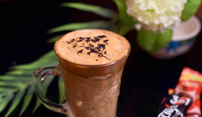 Fotografia de uma xícara de vidro com uma bebida com chocolate, Nescafé e leite. Ao lado, uma embalagem de Nescafé, e ao fundo, decoração de plantas e flores, sobre uma mesa preta.