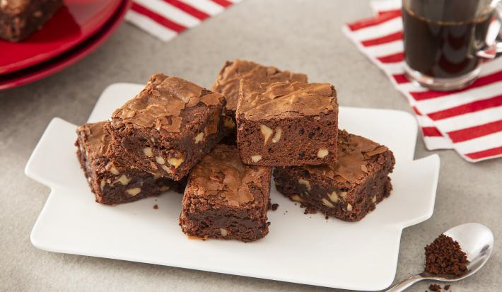 Foto da receita de Brownie de Café. Observa-se 5 pedaços de brownie dispostos um em cima do outro sobre um prato branco. Ao lado direito, uma xícara de café para acompanhar.