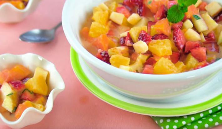 Fotografia em tons de rosa, branco e verde de uma bancada rosa vista de cima, um pano verde com bolinhas brancas e acima um prato branco e verde e por cima do prato um recipiente branco e redondo com frutas. Ao lado um pote branco com frutas.