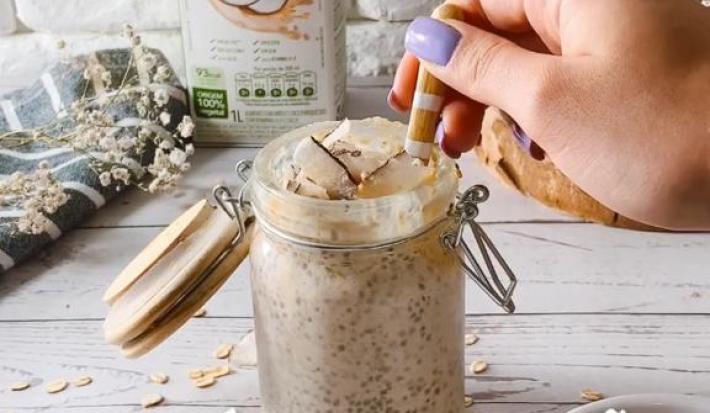 Foto da receita de overnight oats servida em um recipiente hermético alto com uma mão com uma colher, isso sobre uma mesa de madeira cinza. Ao fundo, há uma embalagem de bebida vegetal de coco e arroz antures heart