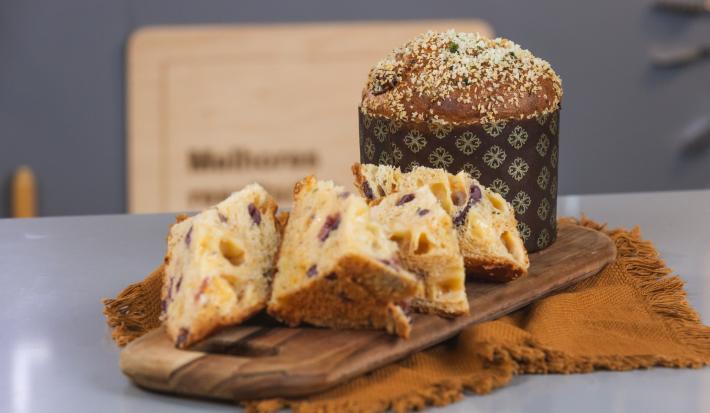 Foto da receita de Panetone Salgado, cortado em fatias, servido sobre uma tábua de madeira, ao fundo há outro Panetone inteiro