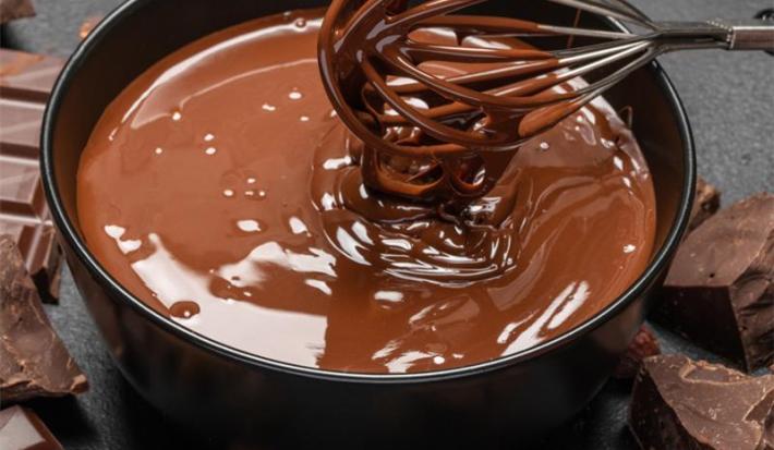 Imagem de um pequeno recipiente contendo uma calda de chocolate cremosa e derretida, em tons de marrom. Um batedor de arame mexe mistura. Ao fundo, barras de chocolate.