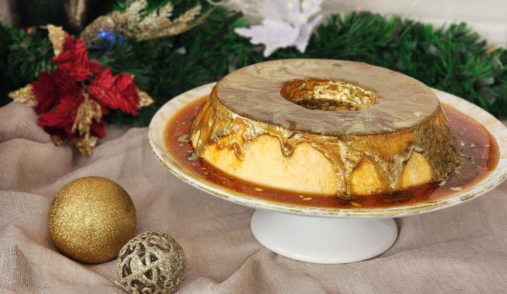 Fotografia em tons de dourado em uma bancada com um pano dourado e enfeites de natal. Ao centro uma bandeja redonda com um pudim de leite Moça e uma calda de caramelo com corante dourado comestível em cima.