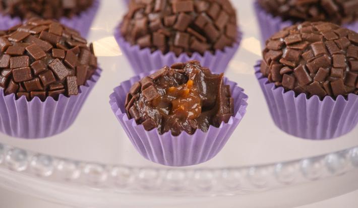 Fotografia em tons de roxo com várias forminhas de brigadeiro roxas ao centro. Dentro de cada forminha existe um brigadeiro de chocolate meio amargo recheado com um creme de caramelo salgado.