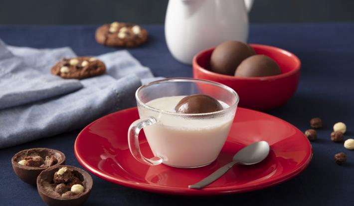 Foto de uma bancada escura. No centro há um prato vermelho com uma xícara com leite no centro e, dentro dela, há uma esfera de chocolate boiando. Ao lado do prato há mais algumas esferas fechadas, uma aberta, alguns cookies e cereal espalhado