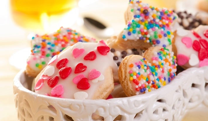 Em cima de uma mesa, tigela com biscoitos redondos e em formato de coração com glacê branco e confeitos coloridos.