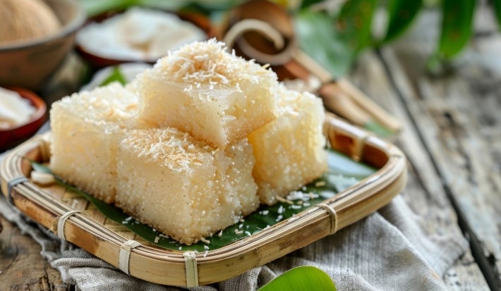 Fotografia de pedaços de bolo de tapioca quadrados dispostos em uma bandeja de bambu forrada com folha de bananeira. O coco ralado no topo e o fundo rústico.