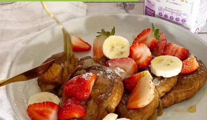 Foto em tons claros da foto da receita de french toast servida com bananas, morangos e uma calda sobre um prato de porcelana branco. Ao fundo, há uma embalagem de bebida vegetal de aveia natures heart