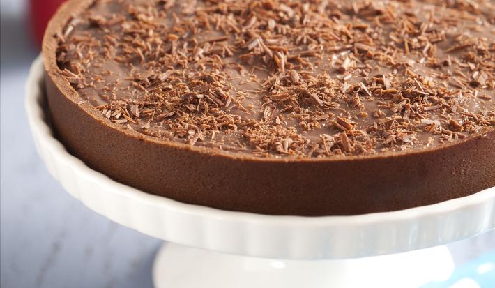 Fotografia em tons de branco e chocolate, ao centro sobre boleira branca uma torta de achocolatado com raspas de chocolate, ao lado guardanapo azul com bolinhas brancas, copo vermelho com talheres e pratos ao fundo, tudo sobre bancada em tons de cinza.