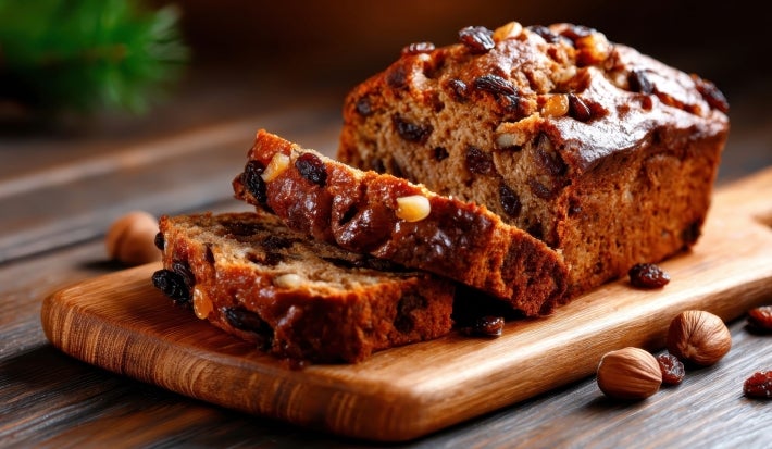 Fotografia de um bolo de frutas e nozes, com crosta dourada e textura rica. Cortado em fatias, revela um interior úmido e denso, repleto de passas e nozes, sobre uma tábua de madeira, rodeado por avelãs e passas.
