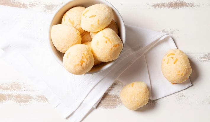 Imagem vista de ima de um bowl branco com pães de queijo. Ao lado, 2 pães de queijo sobre um pano branco.