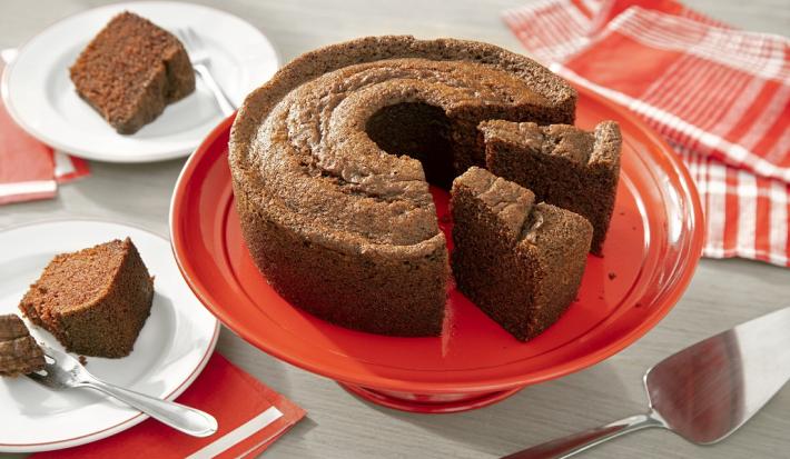 Fotografia em tons de marrom em uma bancada de madeira de cor cinza. Ao centro, uma boleira vermelha contendo o bolo de chocolate. Ao fundo, há um pano vermelho listrado e ao lado, há 2 pires com 2 fatias de bolo.