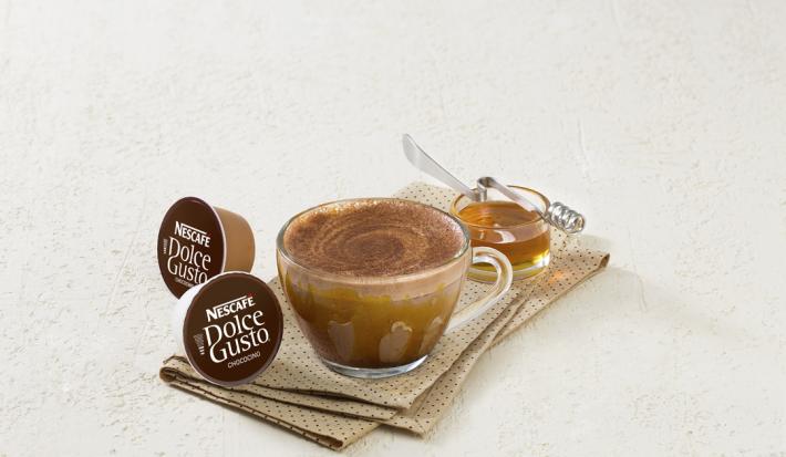 fotografia em tons de branco e marrom de uma bancada branca. Contém um pano marrom com uma xicara transparente com a bebida e duas capsulas de Dolce Gusto ao lado