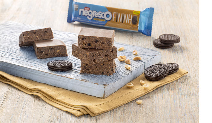 Fotografia mostra quadradinhos de paçoca em tom de chocolate, ao lado amendoins e biscoitos. Ao fundo um pacote de biscoito de amendoim.