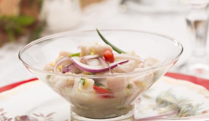 Fotografia em tons de branco e vermelho de uma mesa branca com um prato vermelho, sobre ele um prato branco florido, uma colher prata, e uma taça com pedaços de peixe, cebola roxa, pimenta e cebolinha. Ao fundo um arranjo de flores.
