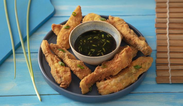 Foto da receita de Costelinha de Tilápia Copacol Empanada com Molho Oriental na Airfryer, servida em um tigela azul escura, com um pote pequeno com o molho no centro do prato, tudo numa bancada azul