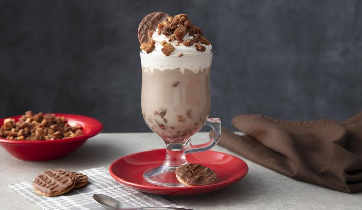 Foto de uma mesa cinza. Ao centro há um prato vermelho com uma caneca alta com a bebida de Chocolate Quente, decorada com Chantilly e o crumble. Na base há um biscoito Calipso e mais dois na mesa. Ao fundo há um prato com mais crumble.