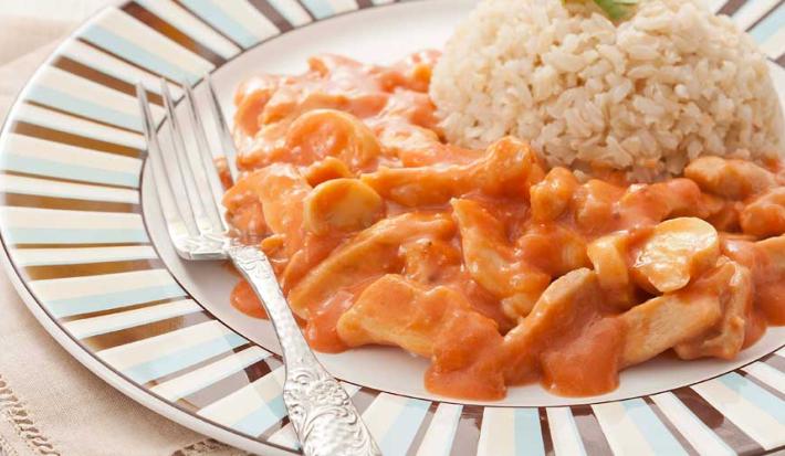 Fotografia em tons de marrom em uma bancada com uma toalha bege, um prato branco raso grande com detalhes listrados em azul e marrom, com o strogonoff de frango com molho e arroz integral.