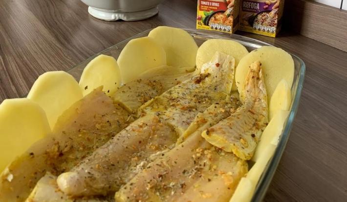 Foto aproximada da receita de Tilápia Assada com Batata no forno, servida em uma travessa de vidro em uma bancada com os temperos maggi meu segredo ao fundo