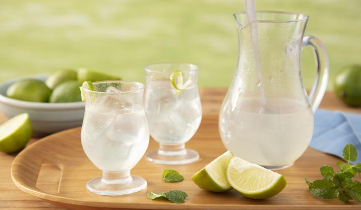 Fotografia em tons de verde em uma bancada de madeira clara com um gramado ao fundo, uma badeja de madeira com uma jarra de vidro com a limonada e dois copos ao lado com a limonada servida, limão e folhas de hortelã espalhadas na bandeja.