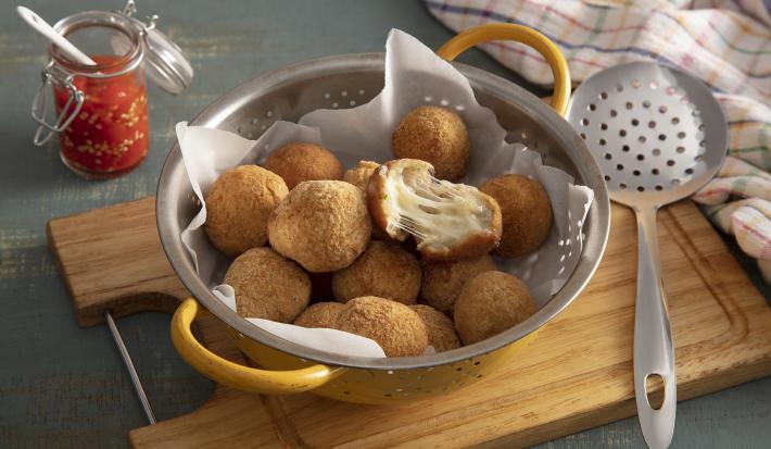 Foto em tons de marrom da receita de bolinho de mandioca (aipim) com queijo servida em diversas porções em um escorredor sobre uma tábua de madeira em cima de uma mesa cinza chumbo. Ao lado há uma escumadeira prateada e um paninho colorido
