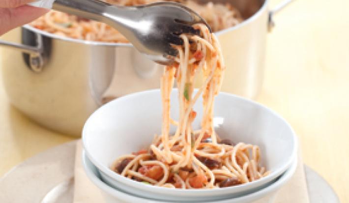 Fotografia em tons de branco e bege, com bowl branco contendo macarrão com molho Putanesca e pegador de massa seguro por uma mão, sobre guardanapo bege e sous plat de inox, panela de inox com macarrão ao lado, tudo sobre bancada bege.