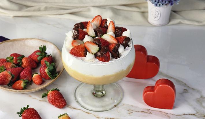 Foto da receita de Morangoffee na Taça, servida em uma grande taça de vidro, em duas camadas, uma de creme bege e outra de creme branco, decorada com morangos com chocolate e suspiros, em uma bancada com mais morangos e alguns corações decorativos