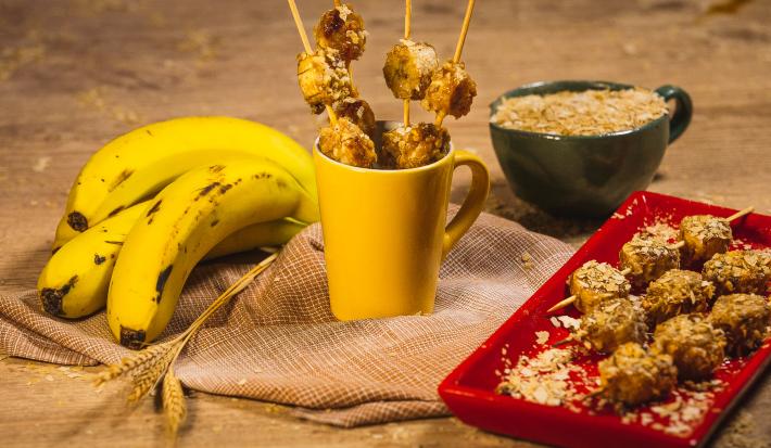 Foto da receita de Banana e Neston no espeto, com alguns espetos dentro de uma caneca amarela e outros em um prato, tudo em uma bancada de madeira decorada com bananas, trigo e um caneca com Neston 3 Cereais dentro.