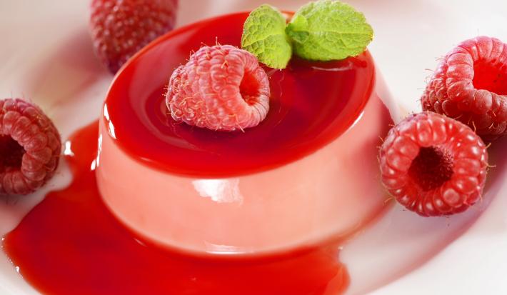 Fotografia em tons de rosa com uma gelatina de frutas vermelhas ao centro. Por cima da gelatina existe algumas framboesas e uma folha de hortelã